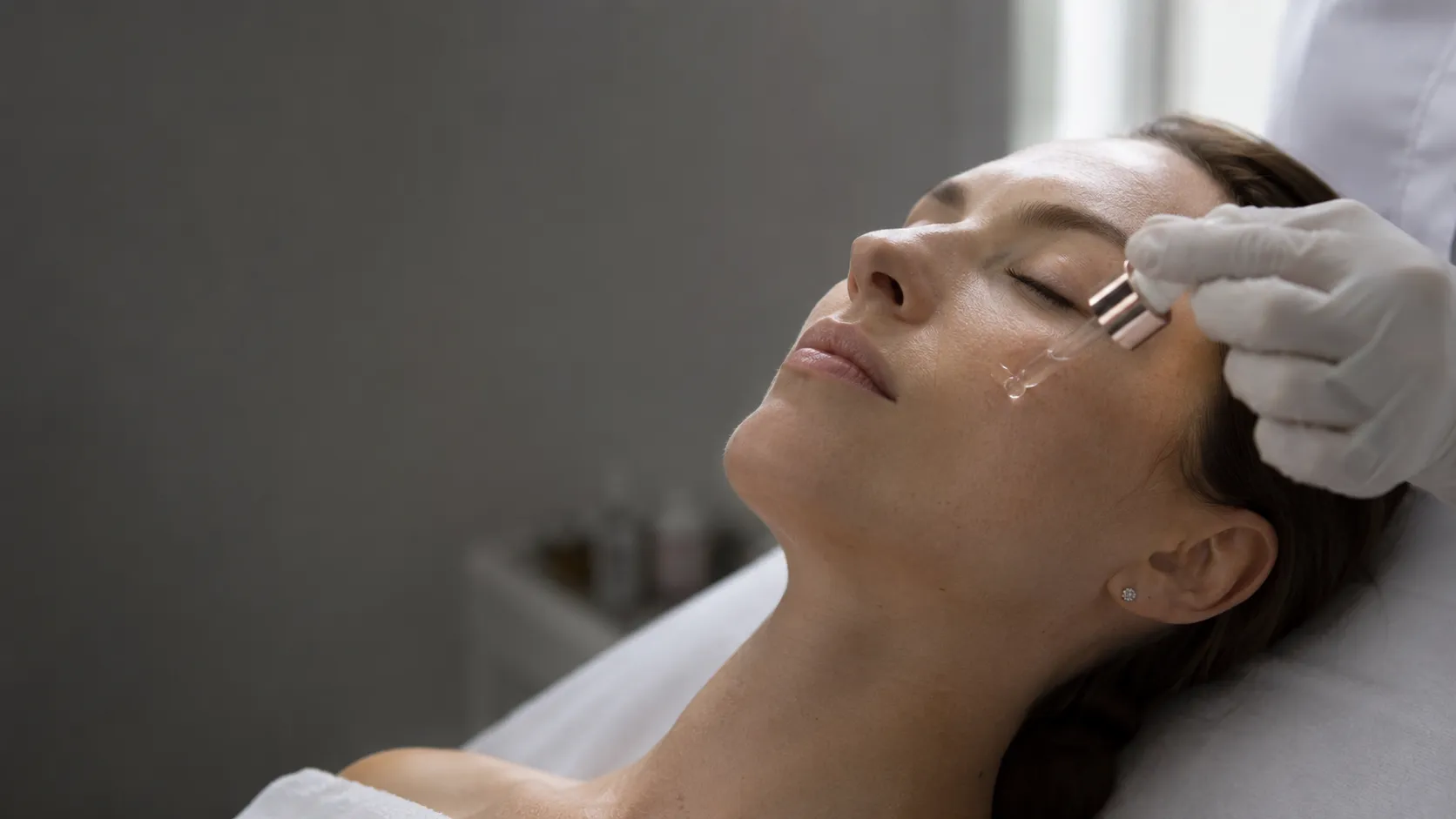Mujer recibiendo un tratamiento facial con sérum aplicado en la mejilla, en camilla de cabina de estética.